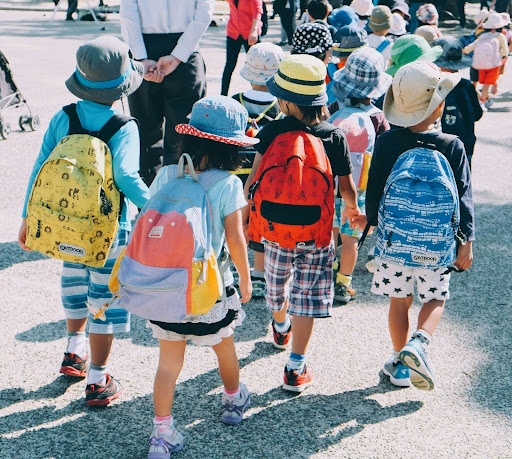 Group of young children wearing backpacks and hats walking together outdoors, representing early childhood routines, transitions, and daily structure.