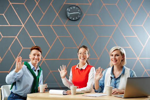 Three professionals sitting at a table, smiling and waving during a meeting, representing teamwork, inclusivity, and positive workplace culture.
