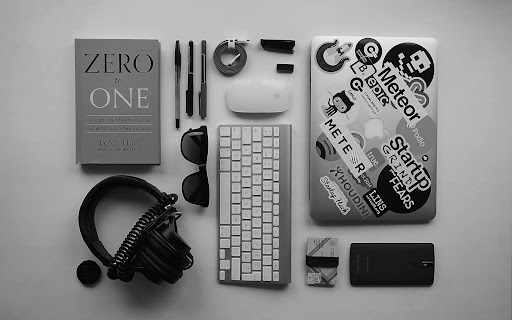 Flat lay of a modern workspace featuring a laptop with stickers, keyboard, headphones, notebook, and everyday professional accessories on a clean desk.