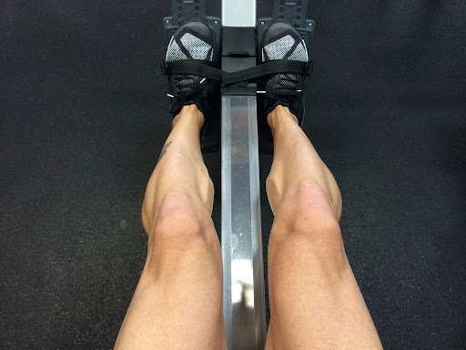 Person using a rowing machine at the gym, showing leg muscles engaged during a fitness and endurance workout.
