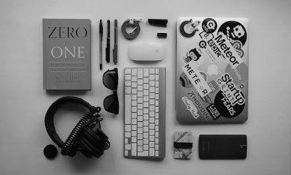 Flat lay of a modern workspace featuring a laptop with stickers, keyboard, headphones, notebook, and everyday professional accessories on a clean desk.