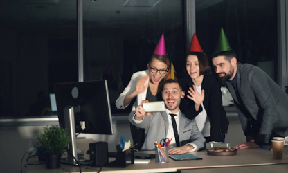 Group of coworkers wearing party hats and smiling around a desk, celebrating a workplace milestone in a modern office environment
