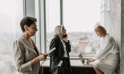 Three women working beside an office window with a notebook, phone and laptop