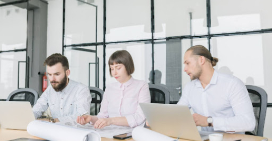 Three professionals working together at a table with laptops and rolled plans, representing teamwork, collaboration, and focused project planning in a modern office environment.