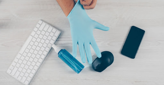 Person putting on protective gloves beside a keyboard, hand sanitiser, smartphone, and mouse, representing hygiene and health awareness in a modern work environment.
