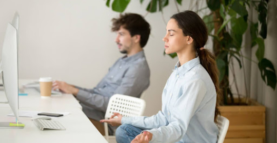 Employee practicing mindfulness meditation at a desk while a colleague works nearby, representing workplace wellbeing, recovery, and mental balance