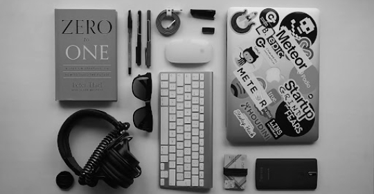 Flat lay of a modern workspace featuring a laptop with stickers, keyboard, headphones, notebook, and everyday professional accessories on a clean desk.