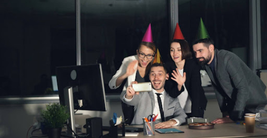 Group of coworkers wearing party hats and smiling around a desk, celebrating a workplace milestone in a modern office environment